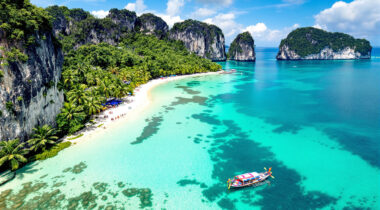 Une plage tropicale thaïlandaise avec un bateau longtail traditionnel naviguant dans des eaux turquoise cristallines près de formations calcaires spectaculaires.